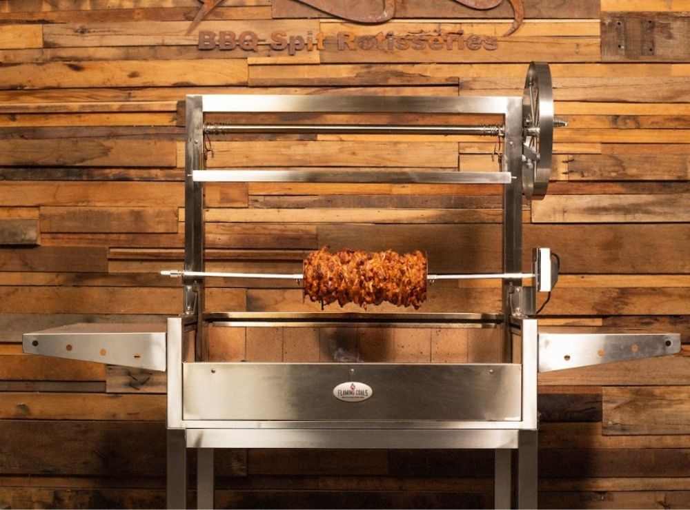 Stainless steel Argentinian Parrilla Grill with a vertical rotisserie attachment, slow-roasting a stacked gyros-style meat skewer over glowing coals.