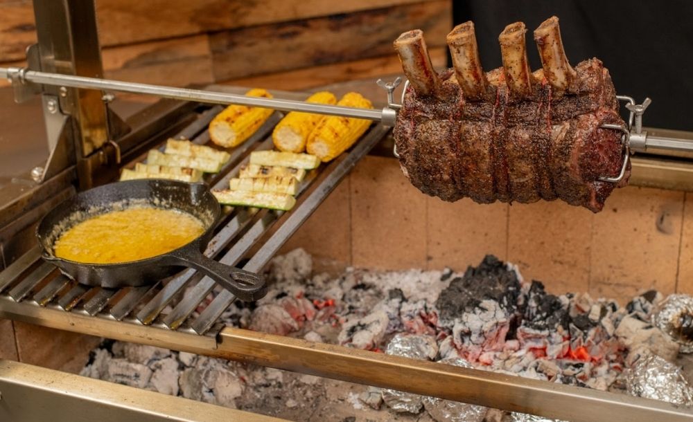 Argentinian Parrilla Grill with a rotisserie ribeye roast turning over hot coals, alongside corn, grilled zucchini, and a cast iron skillet with melted cheese or sauce on the cooking grate.