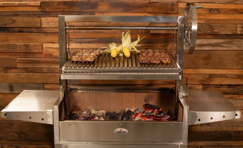 Stainless steel Argentinian Parrilla Grill with adjustable V-grate, cooking steaks and corn over glowing charcoal embers.