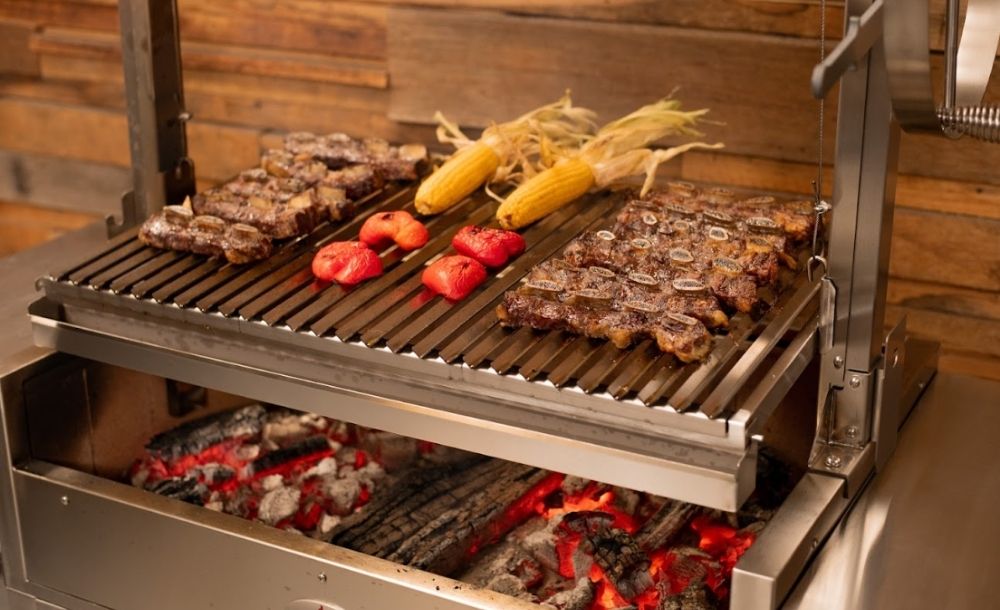 Close-up of a stainless steel Argentinian Parrilla Grill with sizzling meats, corn in husks, and charred red capsicum cooking over glowing charcoal.