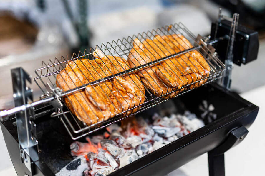 Seasoned chicken pieces cooking in a rotating large multi-use basket over hot charcoal in a spit roaster.