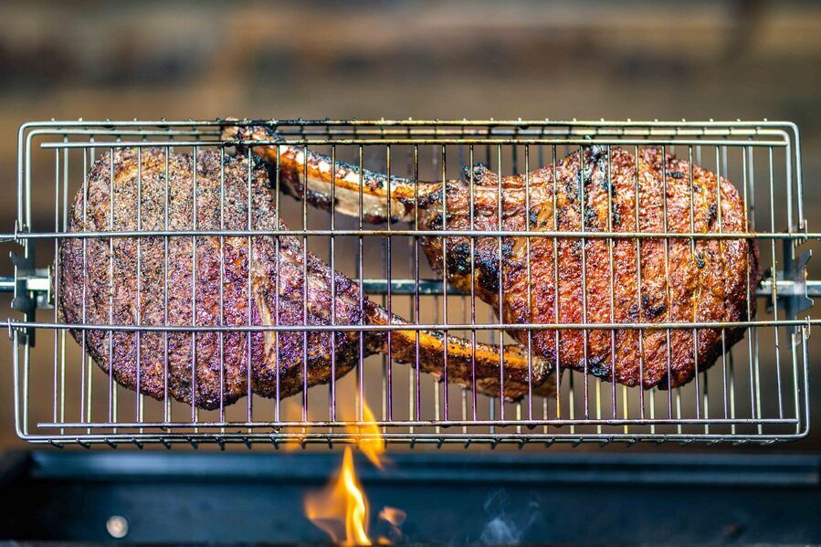 Two large tomahawk steaks cooked in a stainless steel multi-use rotisserie basket over an open flame, showing a charred, caramelised crust.