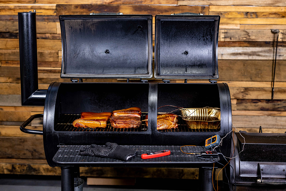  Flaming Coals offset smoker with the lid open, showing several slabs of bacon and pork being smoked alongside foil trays. Temperature probes are inserted in the meat, and BBQ tools are resting on the front shelf.