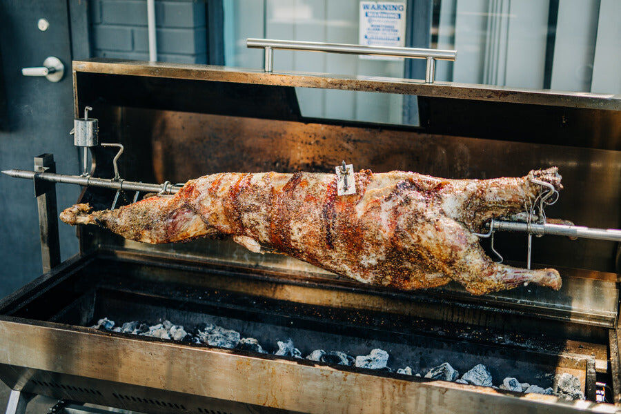 Whole lamb roasting on a commercial spit roaster with a counterbalance weight used to keep the load balanced over charcoal.