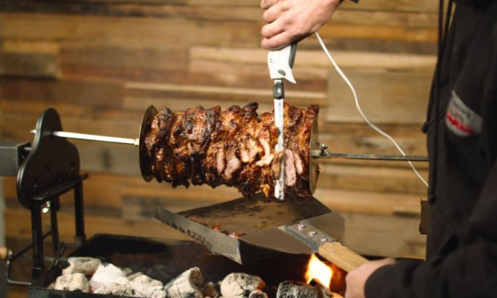 Person carving freshly cooked gyros meat from a rotisserie using an electric knife over hot charcoal, with crispy golden edges visible. Person carving freshly cooked gyros meat from a rotisserie using an electric knife over hot charcoal, with crispy golden edges visible.