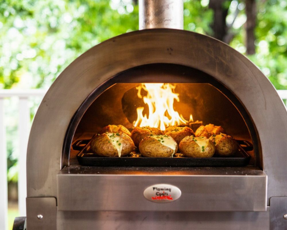 Cheesy baked potatoes cooking inside a Flaming Coals wood-fired pizza oven on a cast iron griddle pan with flames glowing in the background. Cheesy baked potatoes cooking inside a Flaming Coals wood-fired pizza oven on a cast iron griddle pan with flames glowing in the background.