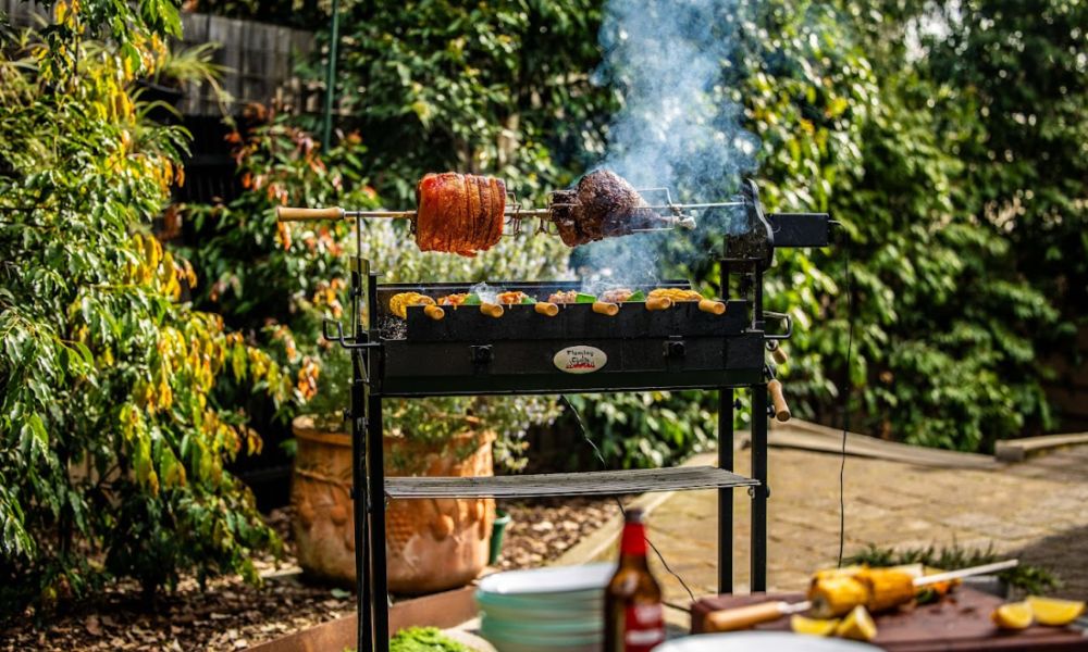 Outdoor scene with a Flaming Coals Cyprus spit rotisserie grilling two large pieces of meat and several skewers of corn over charcoal, with smoke rising against a lush garden backdrop.