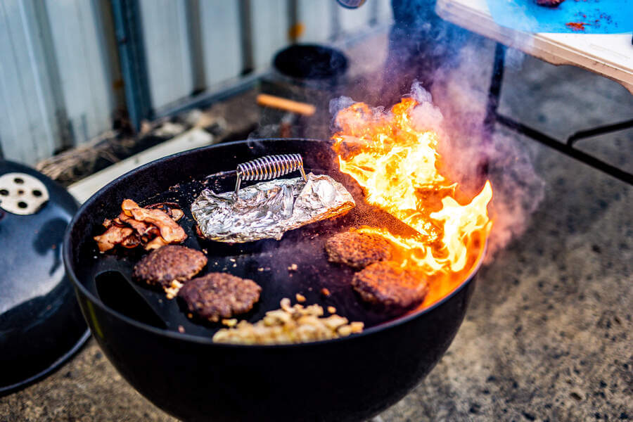 A backyard kettle barbecue with burgers, bacon, onions and a cast iron burger press, with a flare-up flame on the cooking surface