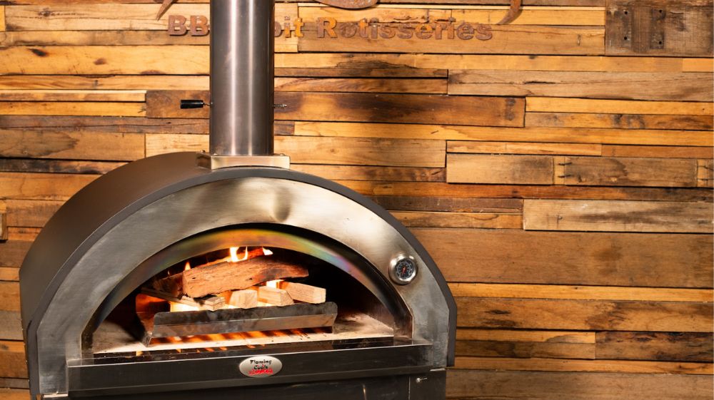 Stainless steel wood-fired pizza oven with chimney, burning logs inside, set against a rustic wooden wall backdrop.