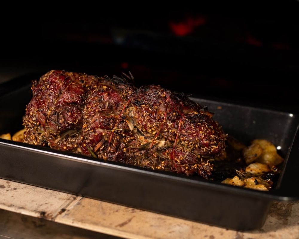 Roast lamb coated in herbs and tied with cooking string, baking in a black tray inside a wood-fired pizza oven with golden potatoes on the side.