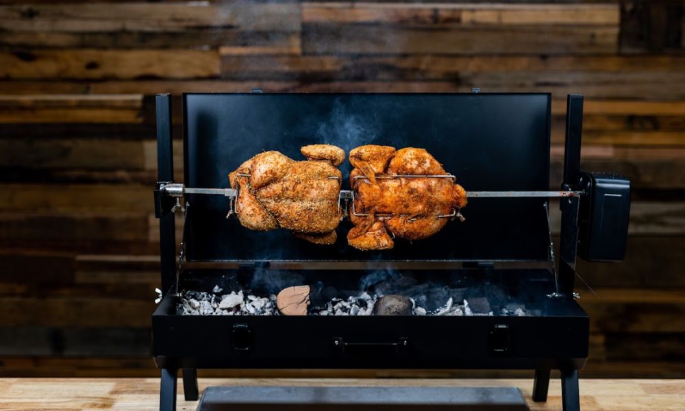 Two whole seasoned chickens cooking on a rotisserie spit over hot charcoal in a black mini spit roaster, with smoke rising against a rustic wooden background.