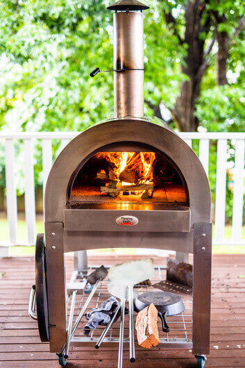 A stainless steel wood-fired pizza oven burning with an active fire, set on a wooden deck with various cooking accessories and firewood stored on the shelf below.
