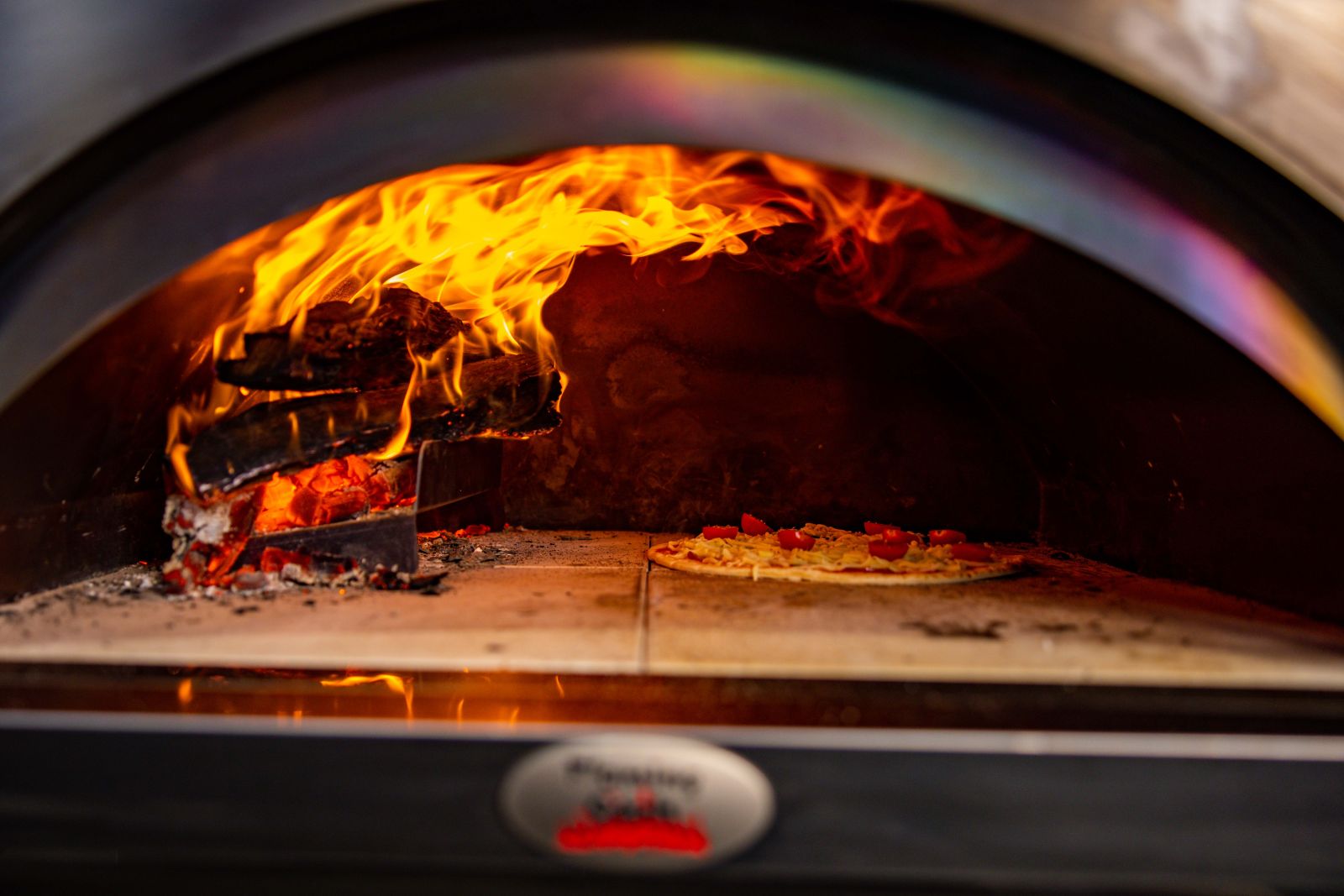 A wood-fired pizza oven with flames burning on one side and a pizza cooking directly on the stone surface inside.