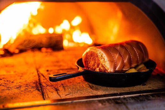 A seasoned roast cooking in a cast iron pan inside a wood-fired pizza oven, with flames burning brightly in the background.