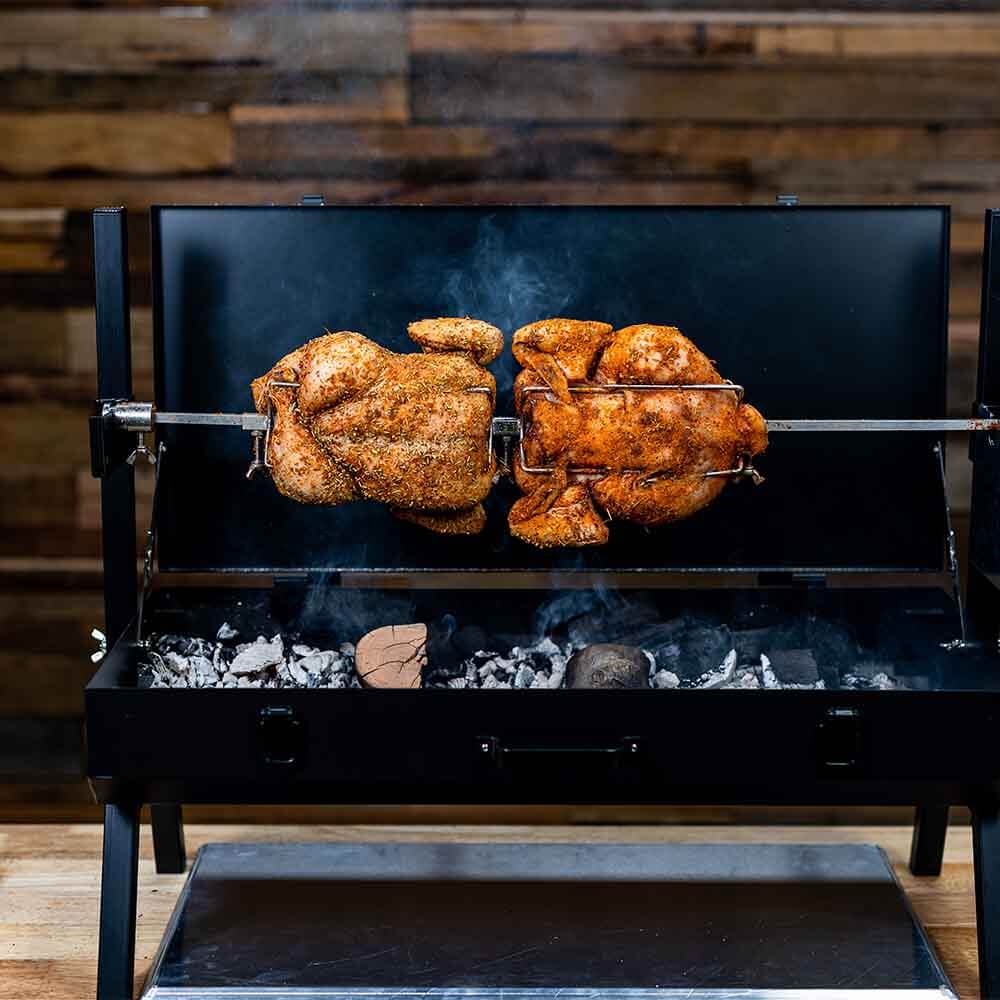 Two seasoned whole chickens roasting on a charcoal spit rotisserie BBQ with smoke rising.
