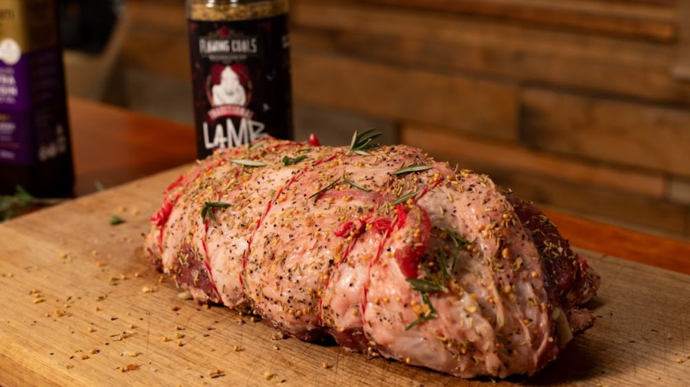 Raw lamb roast tied with string and seasoned with Flaming Coals Lamb Rub and fresh herbs, resting on a wooden chopping board.