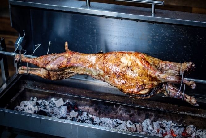 Whole lamb roasting on a spit over hot charcoal in a stainless steel Flaming Coals rotisserie, with crispy golden-brown skin and glowing embers beneath.      