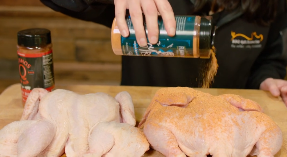 Whole chickens being seasoned with herb and spice BBQ rub on a wooden prep board.