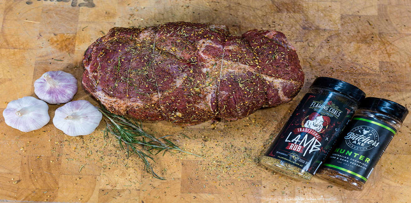 Lamb roast seasoned with rosemary and Greek style spices on a wooden board with BBQ rub containers.