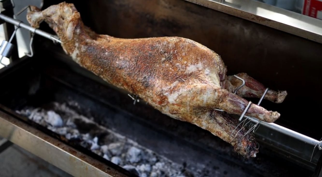 Whole lamb rotating on a spit roaster over hot charcoal during cooking.
