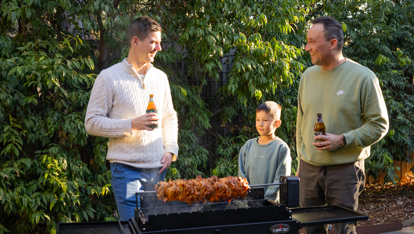 Family gathered around a backyard rotisserie cooking gyros on a charcoal BBQ enjoying outdoor entertaining.