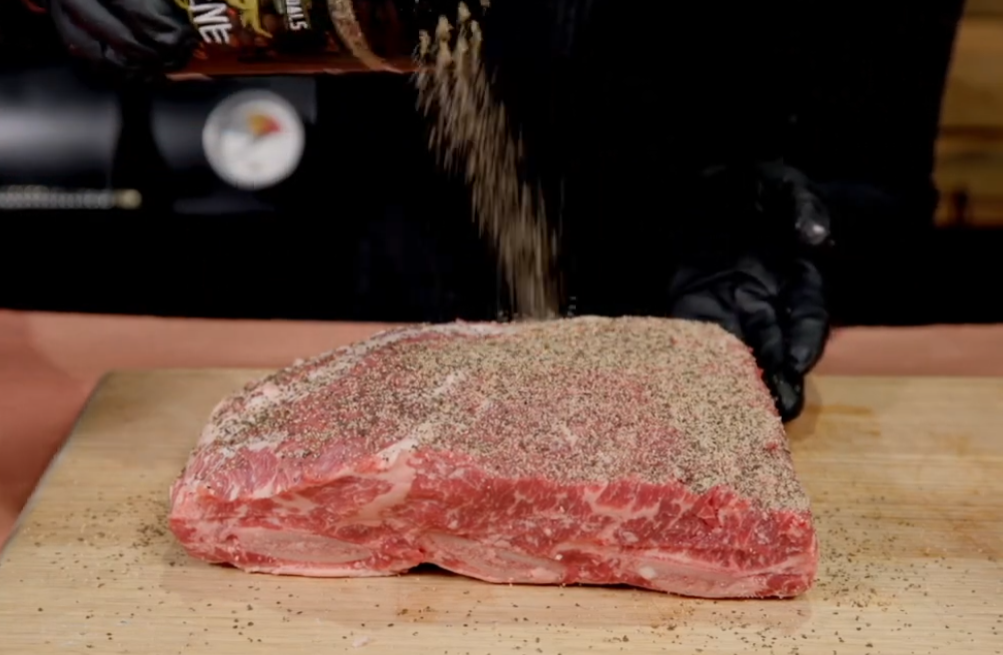 Raw beef brisket being generously seasoned with salt, pepper and spice rub on a wooden board.
