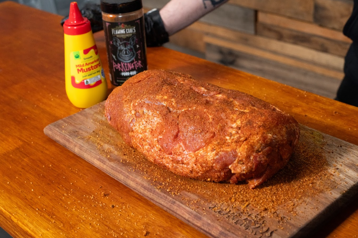 Pork shoulder coated in sweet and savoury BBQ rub on wooden board with mustard and seasoning bottles beside it.