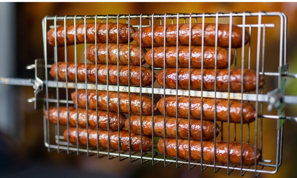 Sausages cooking in a stainless steel rotisserie basket over hot charcoal.